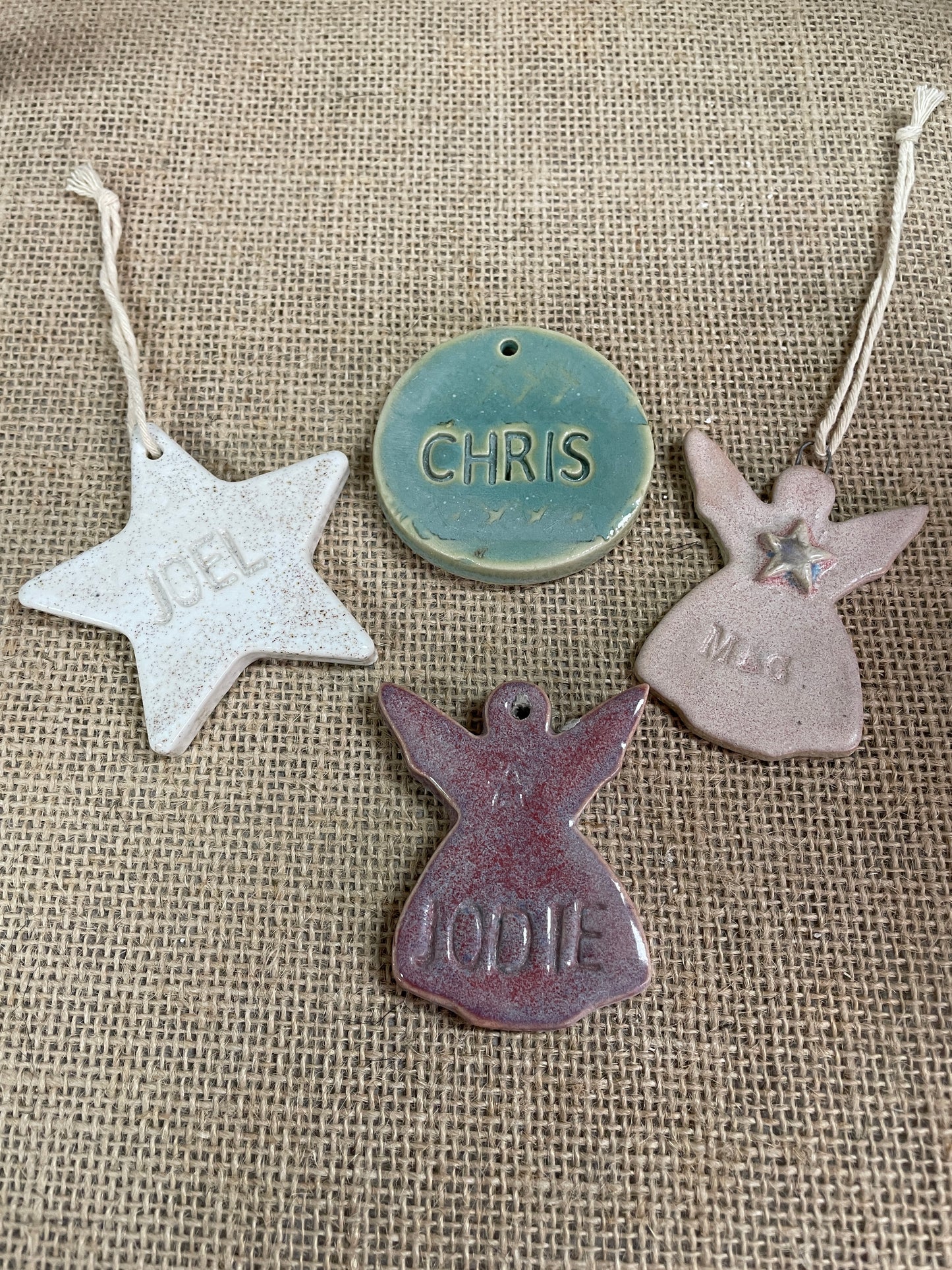 Four personalized ceramic Christmas ornaments in different shapes and colors, including a silver star, a teal round ornament, a pink angel, and a purple angel, displayed on a burlap background.