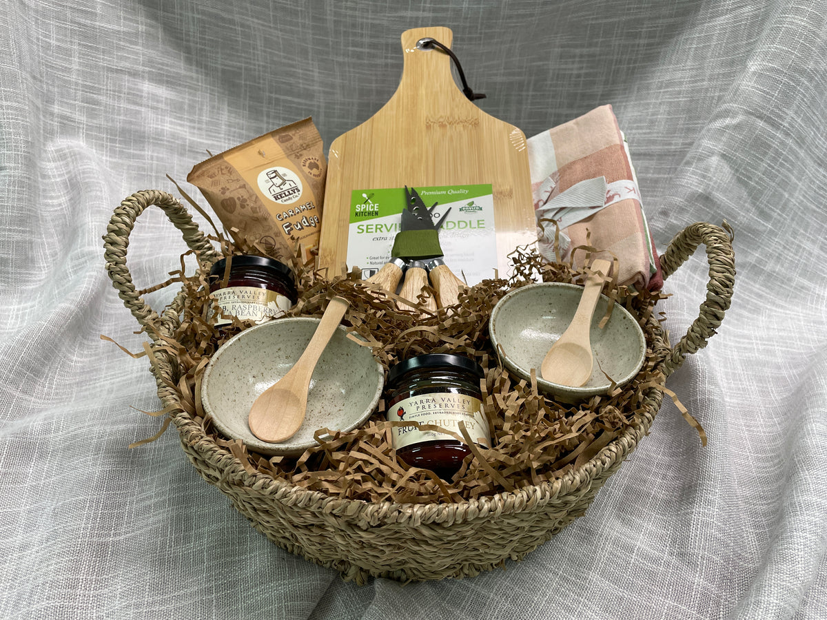 A gourmet gift basket arranged in a woven straw basket with a wooden handle, filled with shredded paper. The basket contains two small speckled ceramic bowls with wooden spoons, two glass jars with dark lids, a wooden cutting board, a bag of coffee, and other packaged food items, all set against a gray textured fabric backdrop.