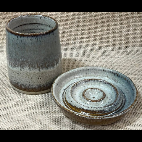 A handmade ceramic cup with a matching saucer lid, featuring a rustic glaze with a gradient from dark brown to light blue-gray, set against a textured burlap background.