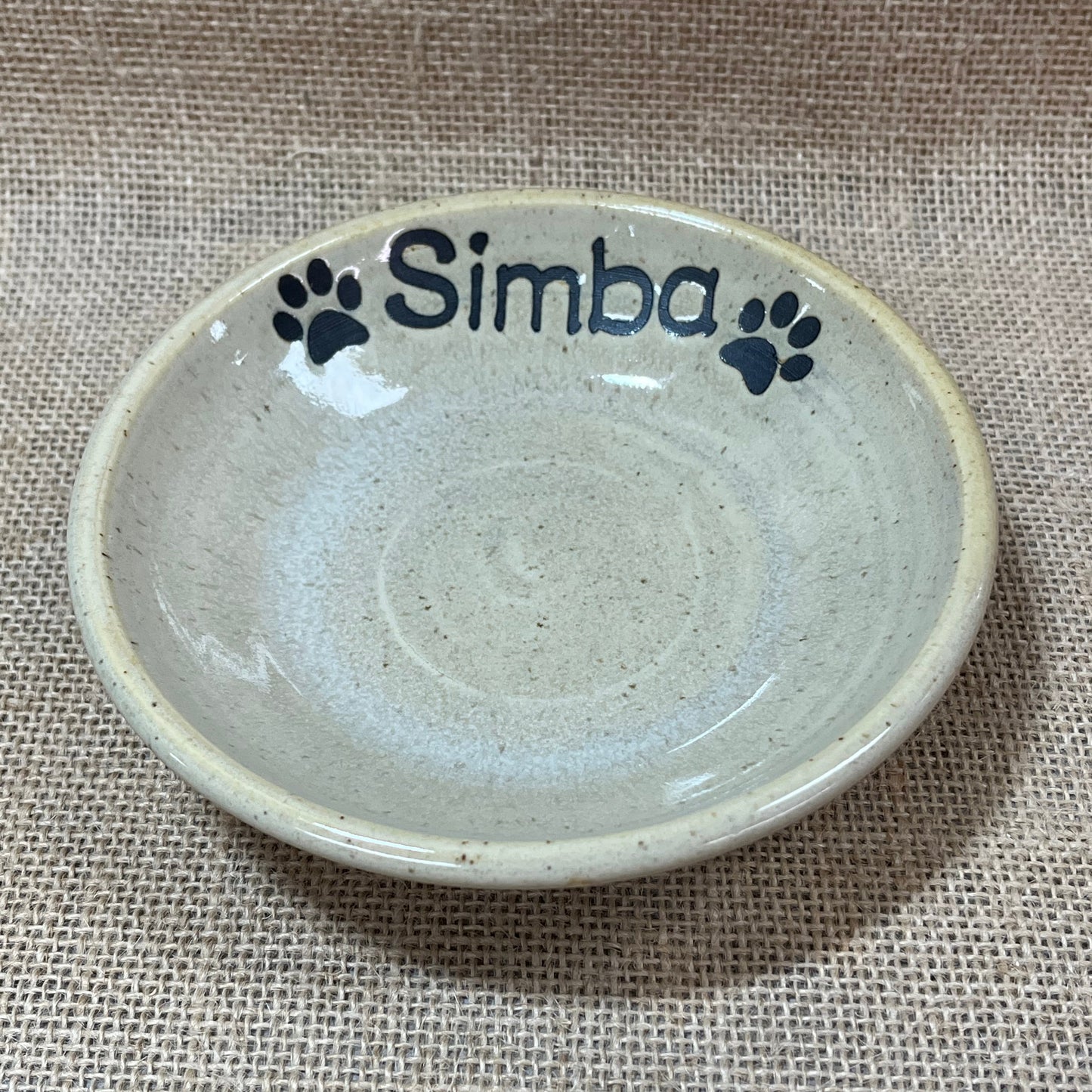 Ceramic pet bowl with 'simba' and paw prints on a textured fabric background
