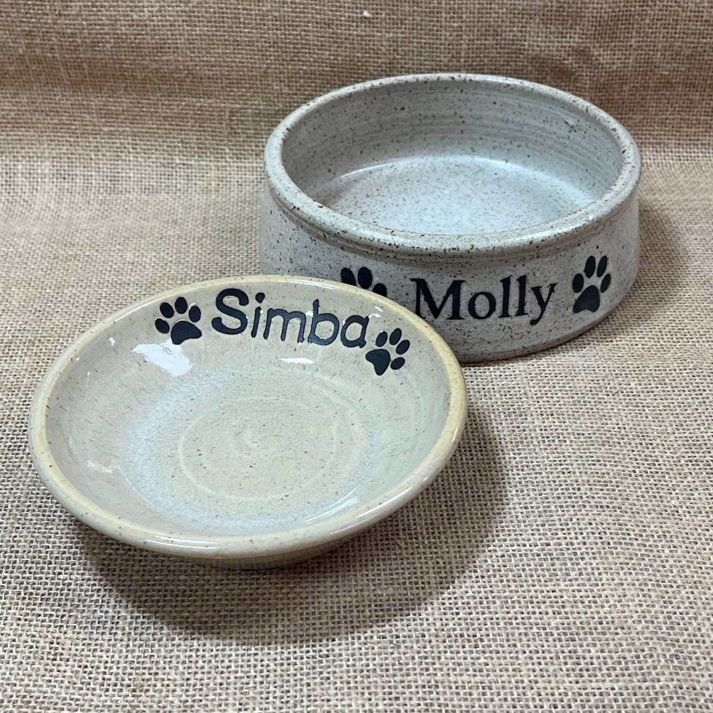 Two ceramic pet bowls with personalized names on a textured fabric background
