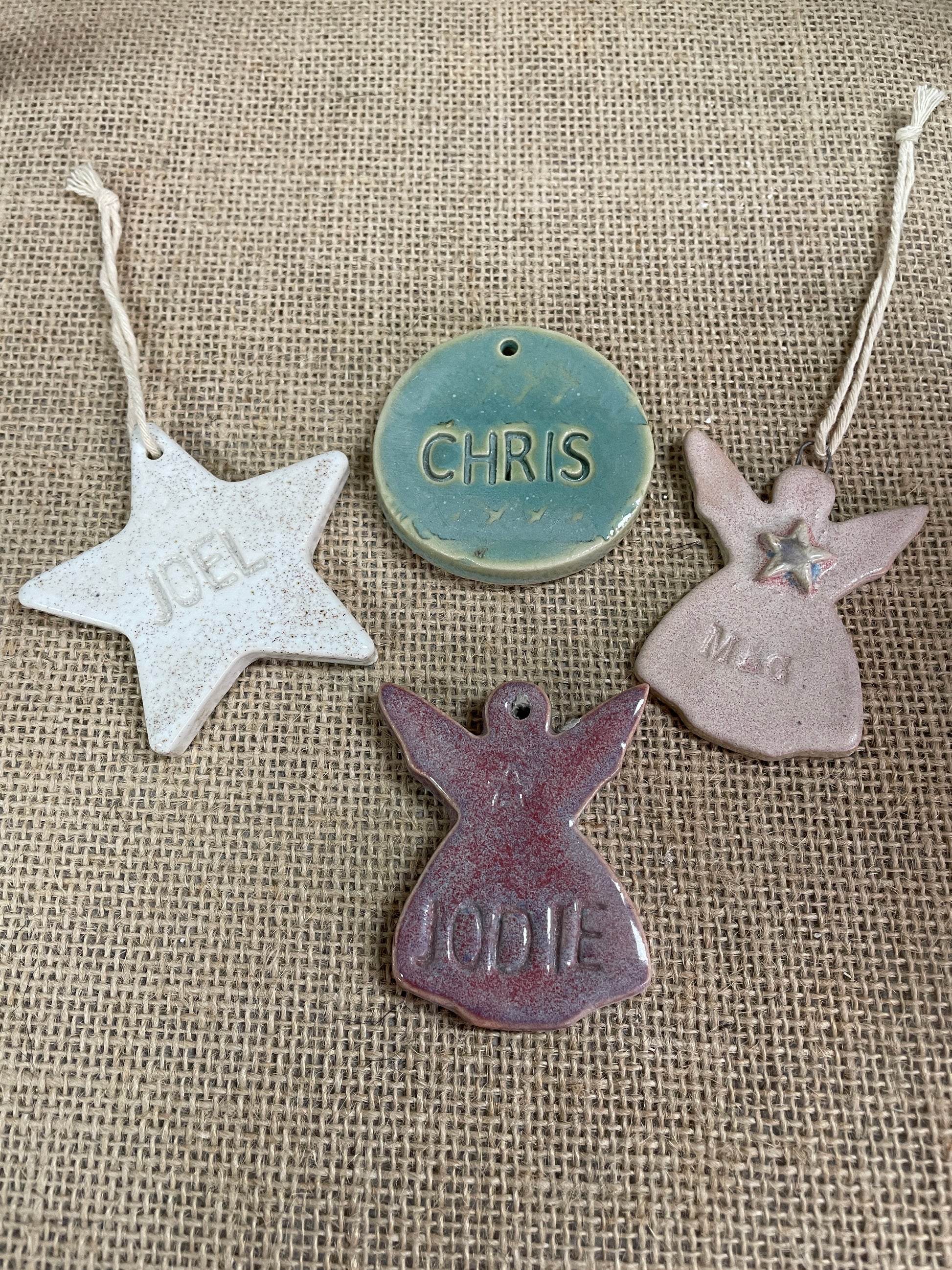 Four personalized ceramic Christmas ornaments in different shapes and colors, including a silver star, a teal round ornament, a pink angel, and a purple angel, displayed on a burlap background.