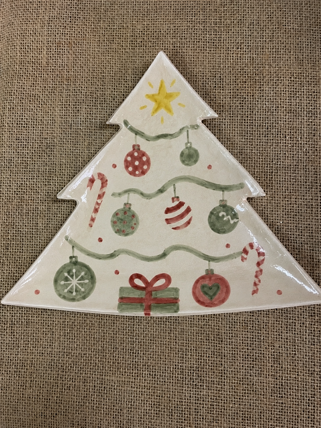 A Christmas tree-shaped ceramic plate with a hand-painted design featuring ornaments, candy canes, a gift box, and a star on a burlap background.