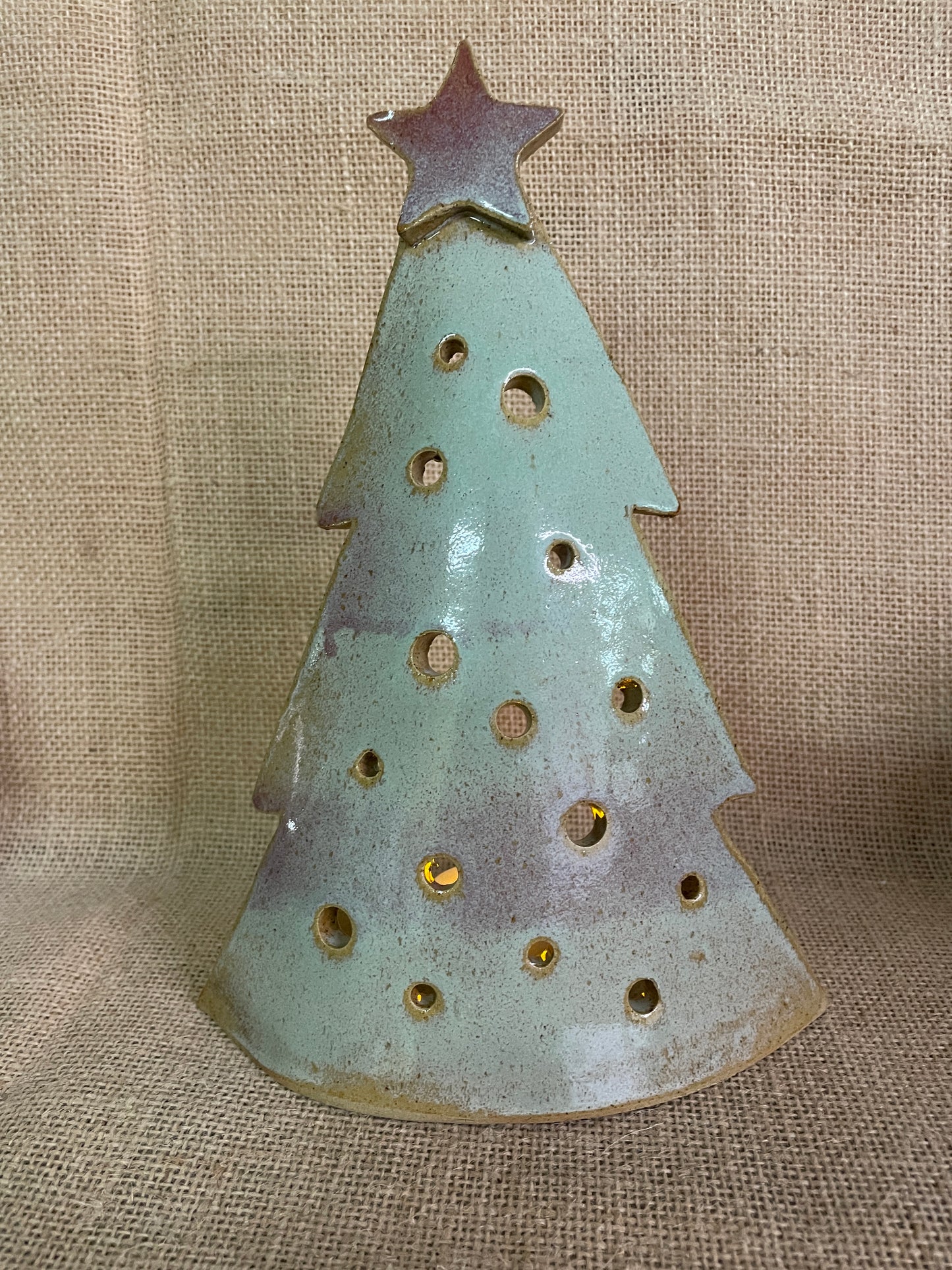 A ceramic Christmas tree silhouette ornament with a star topper, featuring a speckled light blue-green glaze with brown and purple patina accents, set against a burlap background.