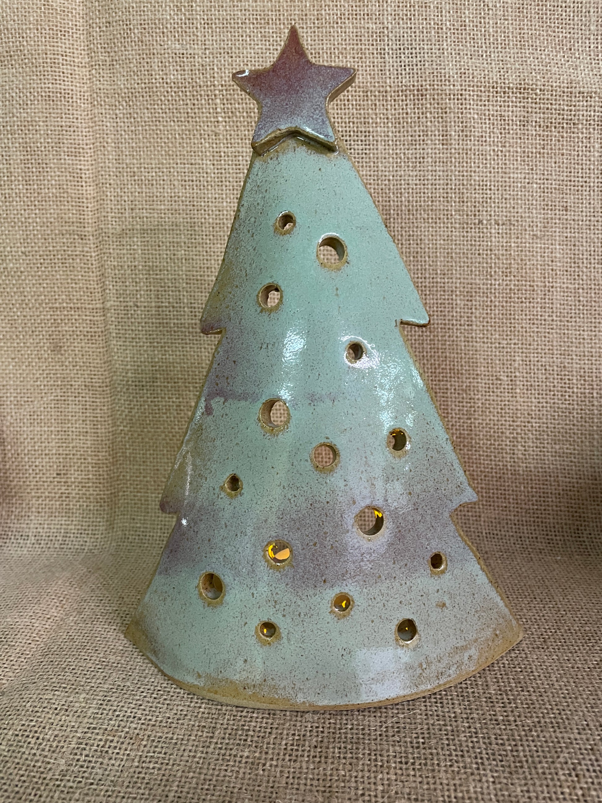 A ceramic Christmas tree silhouette ornament with a star topper, featuring a speckled light blue-green glaze with brown and purple patina accents, set against a burlap background.