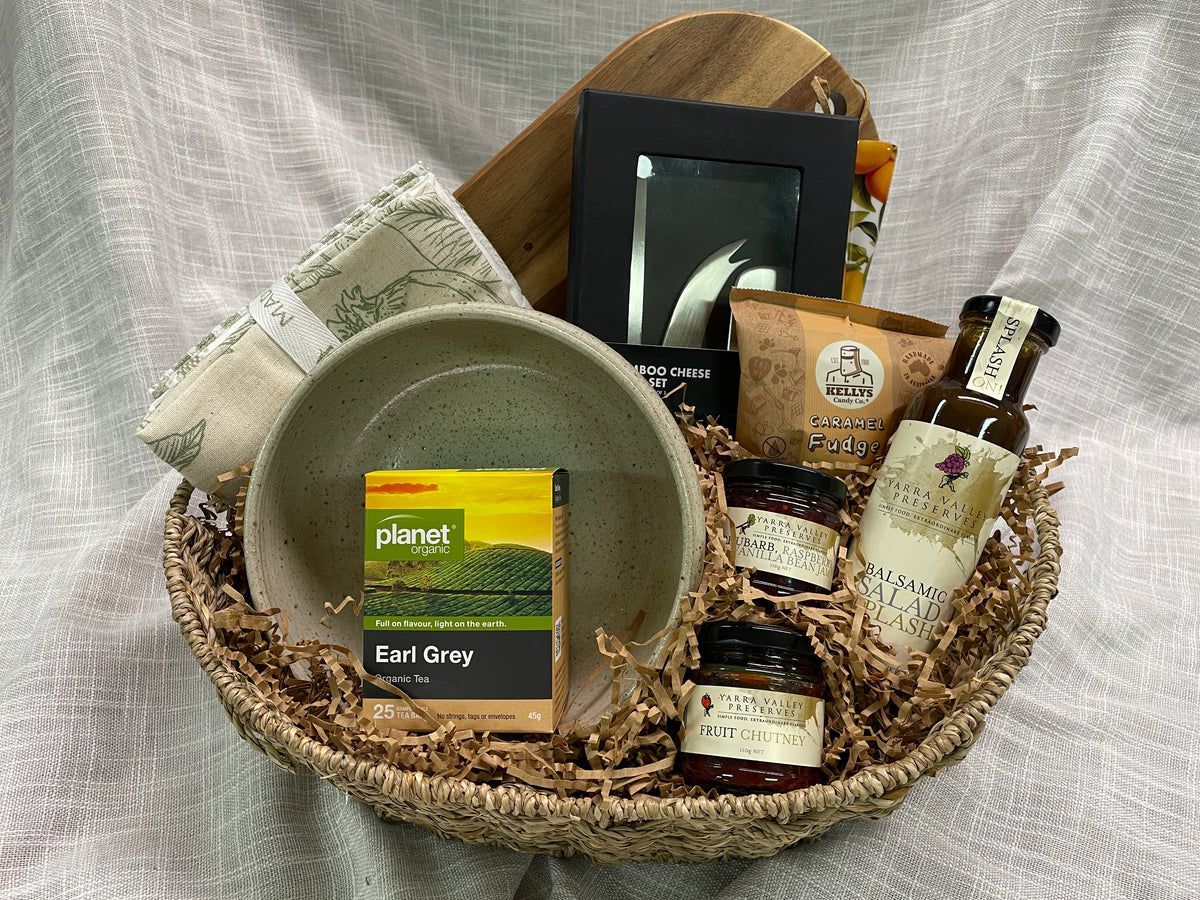 A curated gift basket arranged in a woven basket with shredded paper filler, featuring a speckled ceramic bowl, a box of Planet Earl Grey tea, a bottle of Balsamic Vinegar, jars of preserves, a small framed print, and a folded cloth, all set against a draped light fabric backdrop.