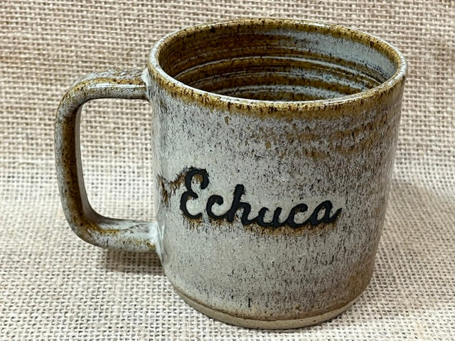 A handmade ceramic mug with a rustic, speckled glaze in beige, brown, and gray tones, featuring the word 'Echuca' written in black script on the front, set against a textured burlap background.