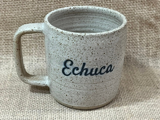 A handmade ceramic mug with a rustic, speckled glaze in white, featuring the word 'Echuca' written in black script on the front, set against a textured burlap background.